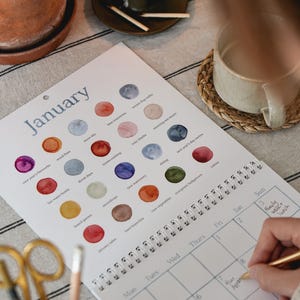 Könnte beinhalten: Ein Wandkalender für Januar mit einem Raster aus Aquarellkreisen, die jeweils mit Texten wie winter dog walks, snow sky und wood fires beschriftet sind. Der Kalender enthält ein Monatsraster mit handschriftlichen Notizen und eine Spiralbindung.