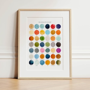 May include: Framed print featuring a grid of watercolor circles in various colors, labeled "the colours of cornwall." The color palette includes shades of blue, orange, green, and brown. The print is in a light wood frame and rests on a wooden surface.