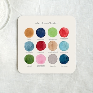 May include: A white coaster with the text "the colours of london." and 16 watercolour circles, each representing a different London landmark. The circles are labelled with the names of the landmarks, such as "Big Ben", "Underground", "Apples and Pears", "Double Decker Buses", "Covent Garden", "Royal Guards", "St Paul's Cathedral", "Camden Market", "Hyde Park", "Columbia Road Flower Market", "Big Smoke", and "River Thames".