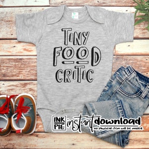 May include: A gray baby onesie with the text "Tiny Food Critic" in black. The onesie is laid out on a wooden surface with a pair of blue jeans and a pair of brown and blue shoes.