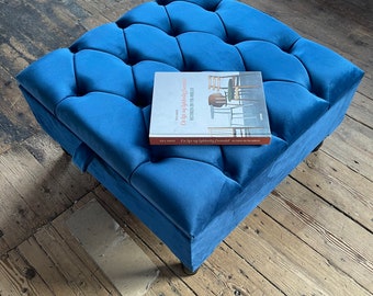 May include: A blue velvet tufted ottoman with a book titled "En lys og lykkelig fremtid" on top. The ottoman has a storage compartment.