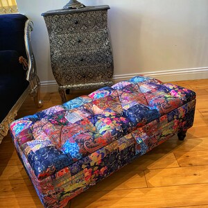 Op de afbeelding: Een kleurrijke patchwork stoffen ottomane met gestoffeerde bekleding en houten poten. De ottomane staat op een houten vloer.