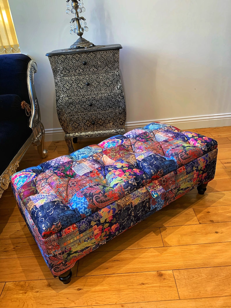 Op de afbeelding: Een kleurrijke patchwork stoffen ottomane met gestoffeerde bekleding en houten poten. De ottomane staat op een houten vloer.