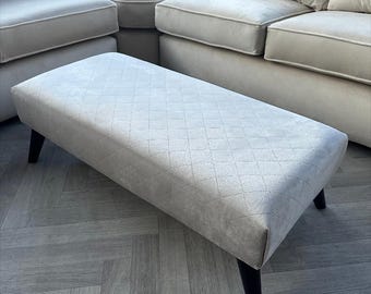 May include: A light gray upholstered ottoman with a diamond-patterned fabric top and black wooden legs. The ottoman is sitting on a light gray wood floor with a herringbone pattern.