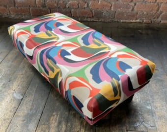 May include: A colorful, abstract patterned footstool with a rectangular shape. The footstool is made of fabric and has a wooden base.