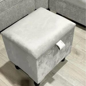 May include: A light grey fabric storage ottoman with a hinged lid and a fabric pull tab. The ottoman has four wooden legs.