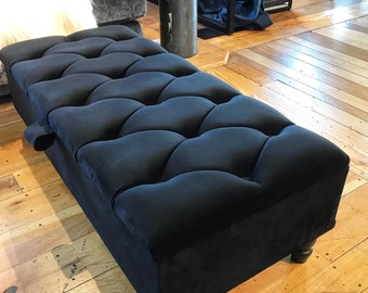 May include: Black velvet storage ottoman with a tufted top and wooden legs. The ottoman is sitting on a wooden floor.