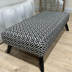 May include: A black and white geometric patterned footstool with black wooden legs. The footstool is sitting on a light wood floor.