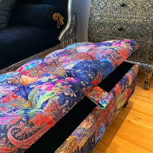 Op de afbeelding: Een kleurrijke, gepatroneerde opbergottoman met een gestoffeerde bovenkant en een zwart interieur. De ottoman is open, waardoor de opbergruimte zichtbaar is.