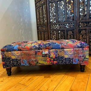 Op de afbeelding: Een kleurrijke patchwork opbergottoman met houten poten. De ottoman staat voor een houten roomdivider met ingewikkelde houtsnijwerk.