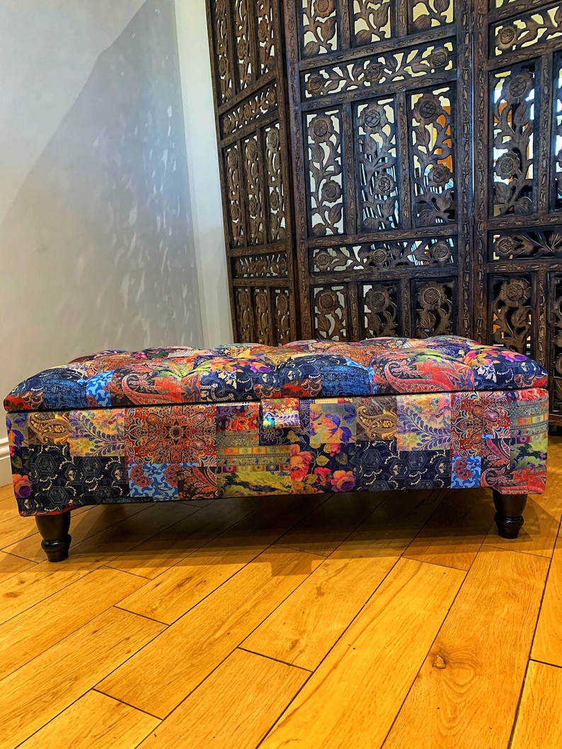 Op de afbeelding: Een kleurrijke patchwork opbergottoman met houten poten. De ottoman staat voor een houten roomdivider met ingewikkelde houtsnijwerk.