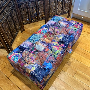 Op de afbeelding: Een kleurrijke patchwork ottomane met een gestoffeerde bovenkant. De ottomane is gemaakt van verschillende stoffen in tinten blauw, roze, groen en paars. De ottomane staat op een houten vloer.