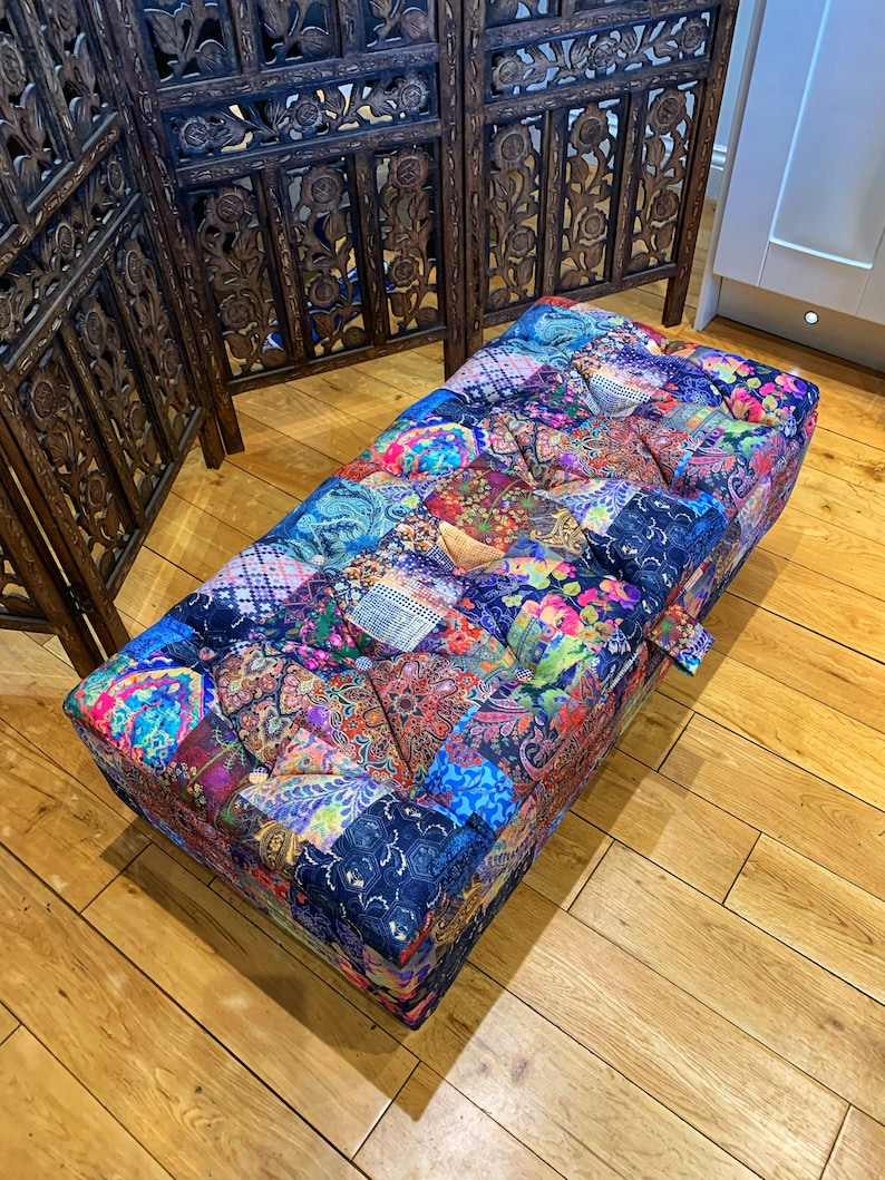 Op de afbeelding: Een kleurrijke patchwork ottomane met een gestoffeerde bovenkant. De ottomane is gemaakt van verschillende stoffen in tinten blauw, roze, groen en paars. De ottomane staat op een houten vloer.