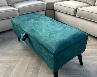 May include: A teal green velvet storage ottoman with black wooden legs. The ottoman has a hinged lid and a handle on the front.