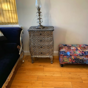 Op de afbeelding: Een zwarte houten dressoir met ingewikkelde houtsnijwerk en een gouden afwerking. De dressoir is bedekt met een decoratieve lamp met een heldere glazen kap en kristallen accenten. De dressoir staat op een houten vloer.