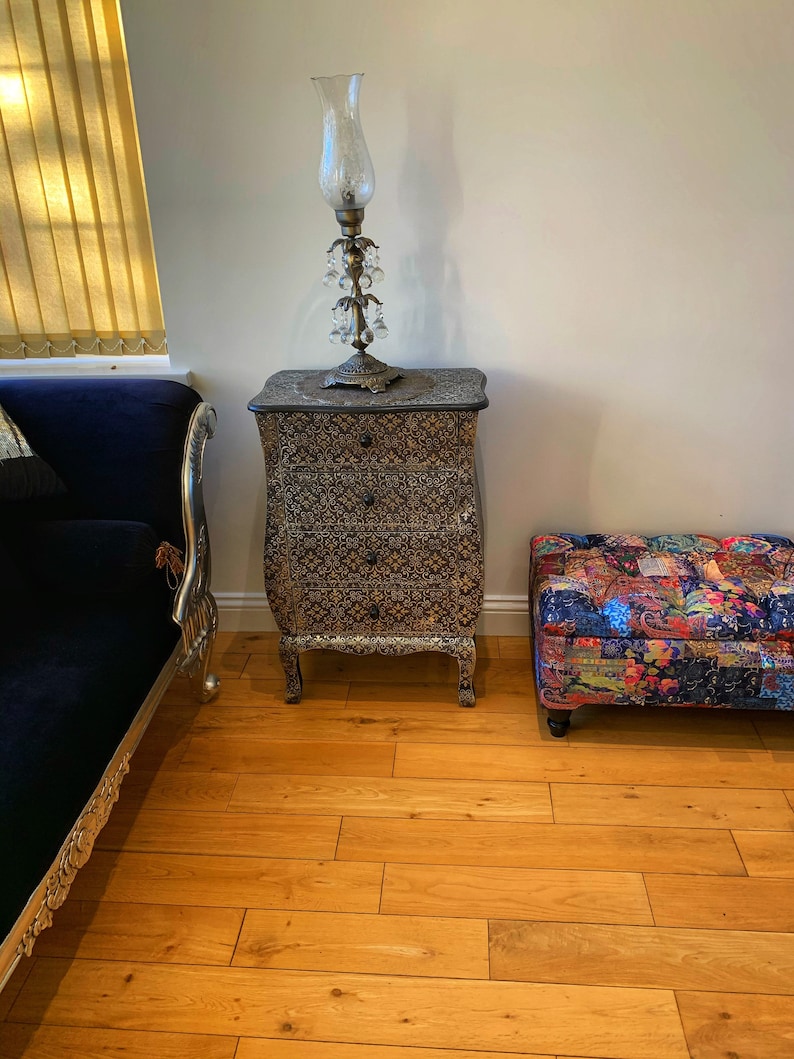 Op de afbeelding: Een zwarte houten dressoir met ingewikkelde houtsnijwerk en een gouden afwerking. De dressoir is bedekt met een decoratieve lamp met een heldere glazen kap en kristallen accenten. De dressoir staat op een houten vloer.