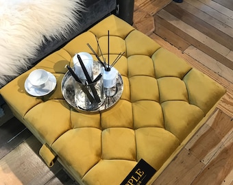 May include: A yellow velvet tufted ottoman with a silver tray on top. The tray contains a white cup and saucer, a silver container, and a glass diffuser with reeds. The ottoman has a black label with the text "RIPPLE INTERIOR" on the side.