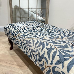 May include: A blue and white floral patterned footstool with wooden legs. The footstool is upholstered in a fabric with a repeating pattern of leaves and branches.
