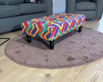 May include: A colorful geometric patterned footstool with black wooden legs. The footstool is sitting on a brown rug.