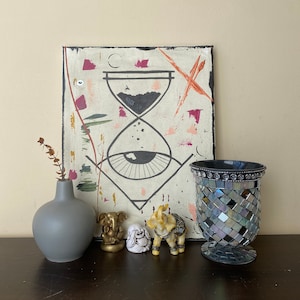Peut inclure: Une peinture en noir et blanc sur toile avec un sablier, un œil et des formes géométriques. La peinture est entourée d'un cadre noir. Il y a trois petites figurines d'éléphants devant la peinture, une dorée, une blanche et une dorée et blanche. Un vase gris et un bougeoir en mosaïque noir et argent sont également présents dans l'image.