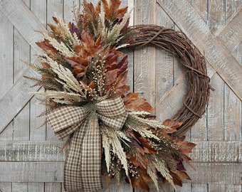 May include: A circular wreath constructed from brown intertwined twigs, decorated on one side with dried brown maple leaves, beige ornamental grasses, and small white berries. A large bow made of brown and beige checkered fabric is attached.