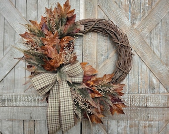 May include: A decorative wreath featuring a circular brown twig base adorned with artificial autumn foliage, including brown maple leaves, green pine needles, and small white berries, finished with a large beige and brown checkered fabric bow.