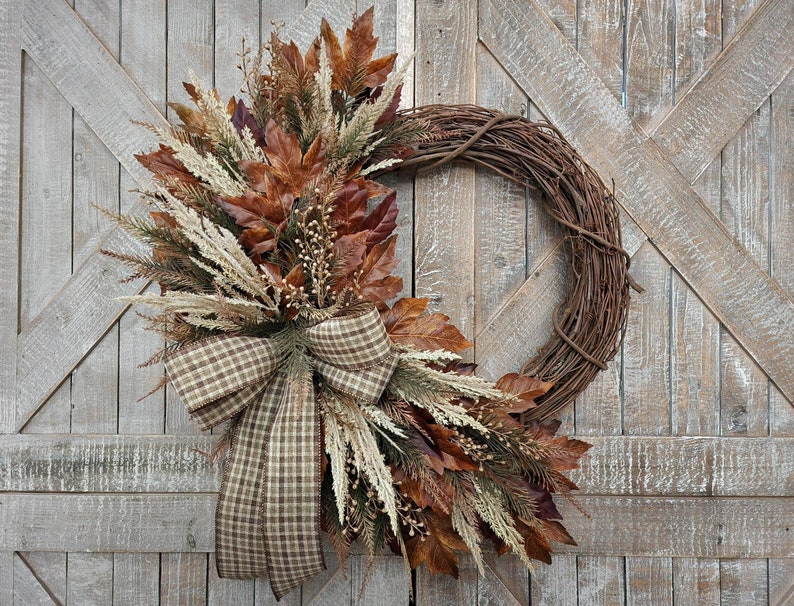 May include: A brown grapevine wreath decorated with fall foliage, including brown and white dried grasses, and a brown and white plaid bow.