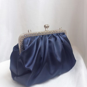 Può includere: Una pochette in raso blu navy con chiusura argentata. La borsa presenta un design arricciato e un aspetto classico ed elegante. La chiusura è un elemento decorativo che aggiunge all'estetica generale dell'accessorio.