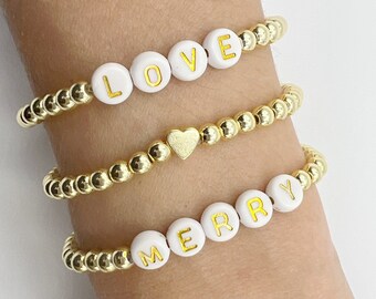 May include: Three gold beaded bracelets with white beads spelling out "LOVE", a heart charm, and "MERRY".