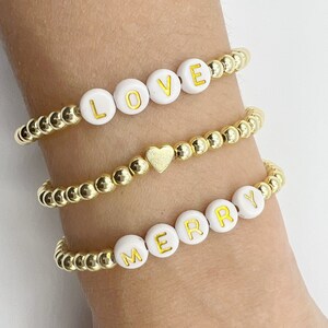 Op de afbeelding: Drie gouden kralenarmbanden met witte kralen die "LOVE", een hartbedel en "MERRY" spellen.