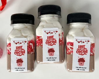 May include: Three clear plastic jars filled with hot cocoa mix, marshmallows, and red and white sprinkles. Each jar has a label that says "Hot Cocoa" with a red heart design. The jars are on a white surface.