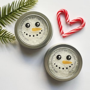 Könnte beinhalten: Zwei silberne Dosen mit Badesalz mit einem Schneemann-Design und dem Text "Dead Sea & Epsom Bath Salts Mix, Christmas Tree Oil, 2 oz". Die Dosen befinden sich auf einer weißen Oberfläche mit zwei roten und weißen Zuckerstangen, die ein Herz bilden.