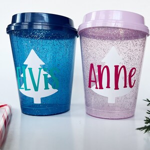 Può includere: Due bicchieri di plastica ricoperti di glitter con coperchi. Uno è blu con "Elvis" in lettere turchesi su un albero di Natale bianco. L'altro è rosa con "Anne" in lettere magenta su un albero bianco. Tema festivo natalizio.