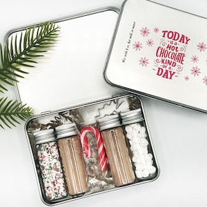 Può includere: Una scatola di metallo argentata con un design rosso e bianco che dice "Today is a Hot Chocolate Kind of Day". La scatola è aperta e contiene quattro piccoli barattoli di vetro con coperchi, una canna di zucchero rossa e bianca e un piccolo tagliapasta a forma di stella bianca. I barattoli contengono codette, miscela di cioccolato caldo, marshmallow e gocce di cioccolato bianco.