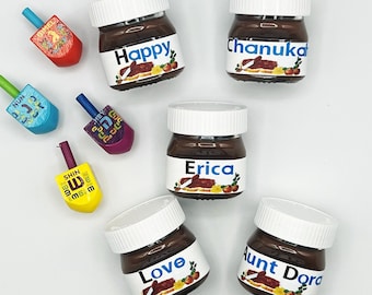 May include: Five small glass jars of chocolate spread with white lids and personalized labels. The labels feature the names "Happy", "Chanukah", "Erica", "Love", and "Aunt Dora".  The jars are surrounded by colorful wooden dreidels.