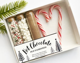 May include: A white box contains a hot chocolate kit with glass tubes of sprinkles, marshmallows, and cocoa powder, plus two candy canes. The box has a label that reads "Hot Chocolate OLD FASHIONED". A sprig of greenery is in the upper left corner.