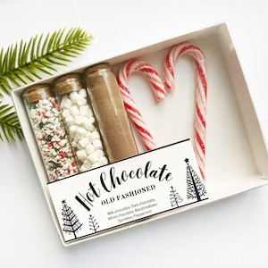 Può includere: Una confezione regalo con un kit per cioccolata calda, tra cui provette di vetro con zuccherini, marshmallow e cacao in polvere, più due bastoncini di zucchero. La scatola ha l'etichetta "Hot Chocolate OLD FASHIONED". Un rametto di verde è in alto a sinistra.