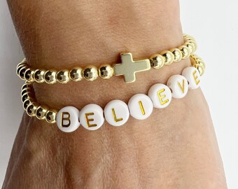 May include: Two gold beaded bracelets, one with the word "BELIEVE" spelled out in white beads with gold lettering, and the other with a gold cross charm.