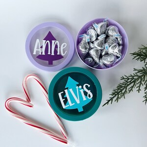 Può includere: Tre contenitori rotondi, due con nomi personalizzati e un disegno di albero di Natale, e uno pieno di caramelle al cioccolato incartate. Un bastoncino di zucchero e un rametto di verde sono anch'essi presenti. I contenitori sono in tonalità viola e verde acqua.
