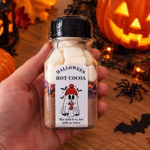 May include: A clear plastic bottle filled with layers of hot cocoa mix, colorful sprinkles, and white marshmallows. The white label features the text HALLOWEEN HOT COCOA, a ghost wearing a red hat, and the instruction Mix with 8 oz. hot milk or water.