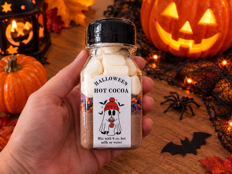 May include: A clear plastic bottle filled with layers of hot cocoa mix, colorful sprinkles, and white marshmallows. The white label features the text HALLOWEEN HOT COCOA, a ghost wearing a red hat, and the instruction Mix with 8 oz. hot milk or water.