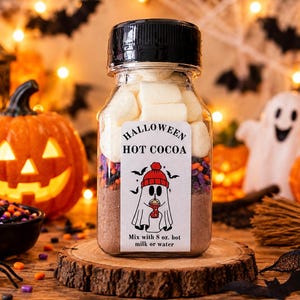 May include: A clear plastic jar filled with layers of hot cocoa mix, colorful sprinkles, and white marshmallows. The label reads HALLOWEEN HOT COCOA with a ghost illustration and instructions to mix with 8 oz. of hot milk or water.