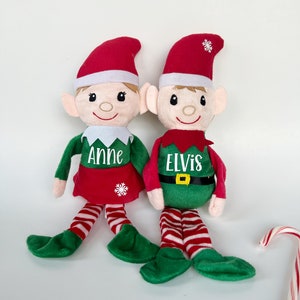 May include: Two plush elf dolls with red hats and green outfits. The elf on the left has the name "Anne" embroidered on its shirt and the elf on the right has the name "Elvis" embroidered on its shirt.