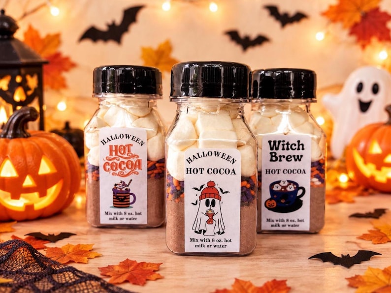 May include: Three clear plastic jars filled with hot cocoa mix, colorful sprinkles, and white marshmallows. Each jar has a black screw-top lid and a white label featuring Halloween-themed graphics and text that reads Mix with 8 oz. hot milk or water.