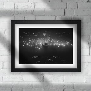 May include: Black and white photograph of a concert with a person silhouetted in the center raising their arm in the air. The person is on a stage in front of a large crowd of people holding up their phones with flashlights on.
