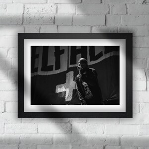 May include: Black and white photo of a musician singing into a microphone on stage. The stage has a large banner with the word "ELF" on it.