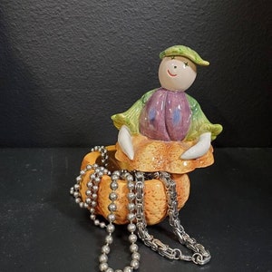 May include: A ceramic pumpkin-shaped trinket box with a smiling character figurine inside. The box is open and two silver chains are spilling out.