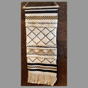 May include: A white and black woven wall hanging with a geometric pattern and gold accents. The hanging has a fringe border and is mounted on a wooden dowel.