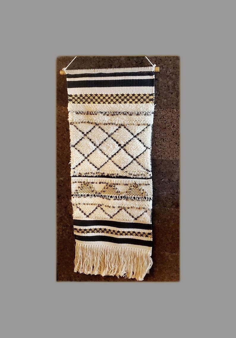 May include: A white and black woven wall hanging with a geometric pattern and gold accents. The hanging has a fringe border and is mounted on a wooden dowel.