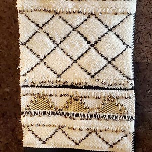 May include: A white and black woven wall hanging with a fringe border. The hanging features a geometric pattern with black diamonds on a white background. There are also gold accents throughout the design.